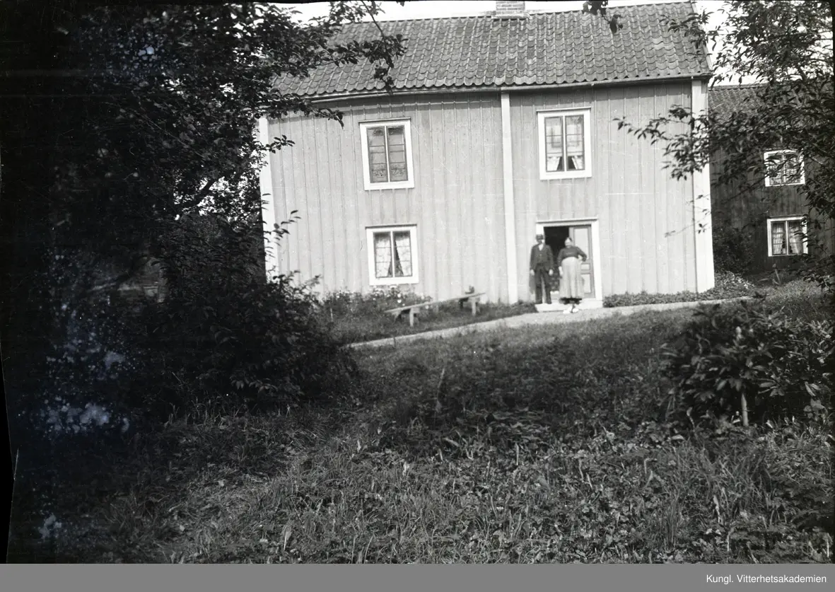 En man och Ketty Andersson framför dörren i Kettys hus i Stensjö by. Huset ör en ljus byggnad i två våningar med enkupigt tegeltak. I fönstrarna hänger gardiner. Till vänster om dörren syns en bänk. Framför huset löper en grusgång. I förgrunden på sidorna syns fruktträd och pion.