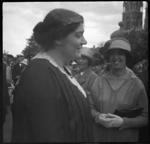 Elsa Warburgs disputation. Kvinnor framför universitetshuset i Uppsala. Warburg i förgrunden bär en lagerkrans. I bakgrunden Uppsala domkyrka.