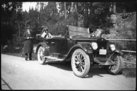 Två kvinnor sitter i en bil på en väg. Bakom bilen står en man, troligen Gösta Laurell. I bakgrunden skog.