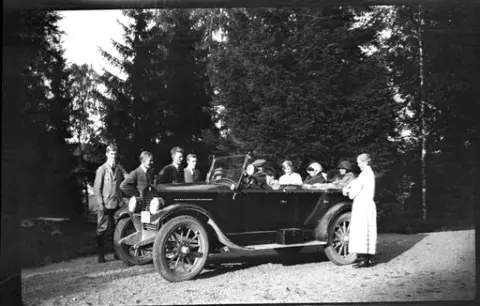 Tretton personer i och runt en bil på en väg. I framsätet sitter tre barn med skärmmössor.