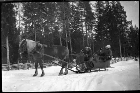 En man och två kvinnor i en släde flätad i korg dragen av en häst på snötäckt mark. Hästen har bjällror monterade på seldonet. Mannen sitter längst fram och håller i tömmarna. Han bär en päls och bredvid honom är en piska. Kvinnorna har fällar över knäna. I bakgrunden syns en gärdesgård i trä och skog.