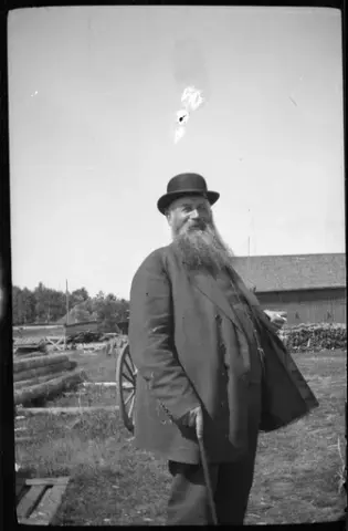 En man i plommonstop och stort skägg håller i en käpp och en cigarett. I bakgrunden syns en vagn, en byggnad med spåntak och flera upplag med stockar och träkonstruktioner.