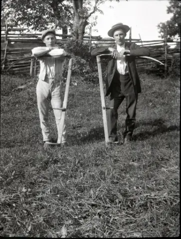 Eugén Stensiö och en ung man med varsin lie i en nyslagen äng. Eugén bär ljust randig väst med matchande byxor, vit skjorta och skärmmössa. Den andra mannen bär en stråhatt och mörka kläder med vit skjorta. Liebladen är fästa med bundna vidjor på orven. Även knaggarnas fäste syns tydligt. I bakgruden syns en gärdesgård i trä bunden med vidjor och en hamlad ask.