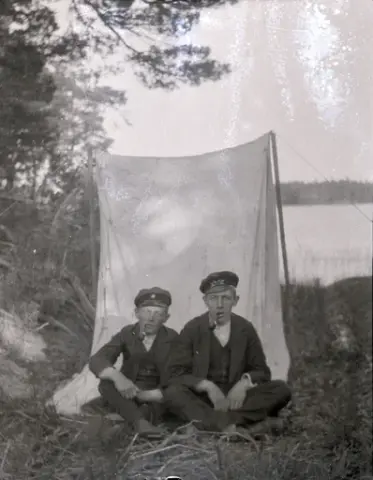 Två pojkar röker pipa framför ett tält. Till höger är Bengt Stensiö. I bakgrunden syns en sjö och skog.