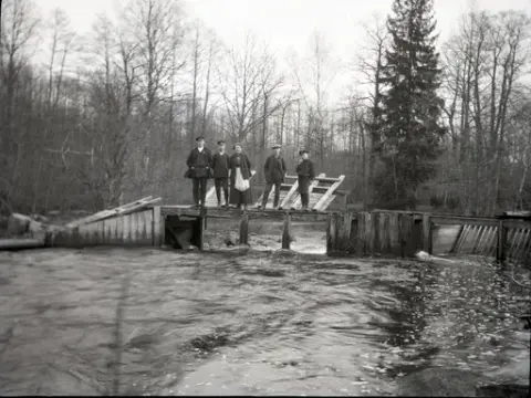 Ett strömt vattendrag med en öppen fördämning byggd i trä. Mot ett räcke står flera dammluckor i trä. Framför dem står fyra pojkar och en flicka. Andra från vänster är Bengt Stensiö, därefter Ingrid Erlandsson och Eugén Stensiö. Pojken längst till höger röker pipa. I bakgrunden syns tät lövskog med inslag av gran.