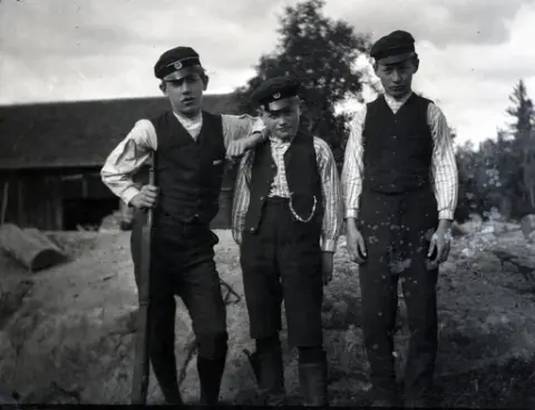 Tre pojkar i Stensjö by. Till vänster är Bengt Stensiö med gevär i handen. Till höger står Eugén Stensiö. I bakgruden syns en lada.