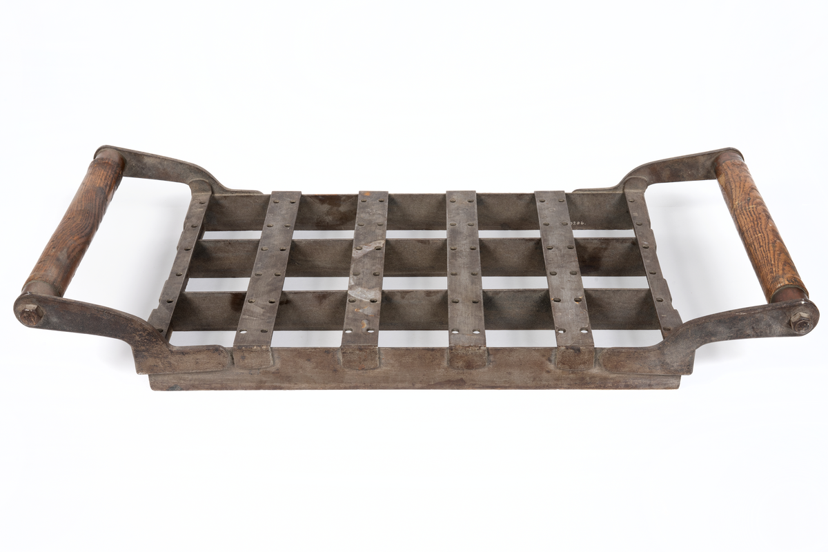 Rektangulär form av järn med två trähandtag längs kortsidorna. Formen är indelad i 15 lika stora rutor. Använd för bakning av skeppsskorpor så kallade knax vid Kronobageriet i Karlskrona.