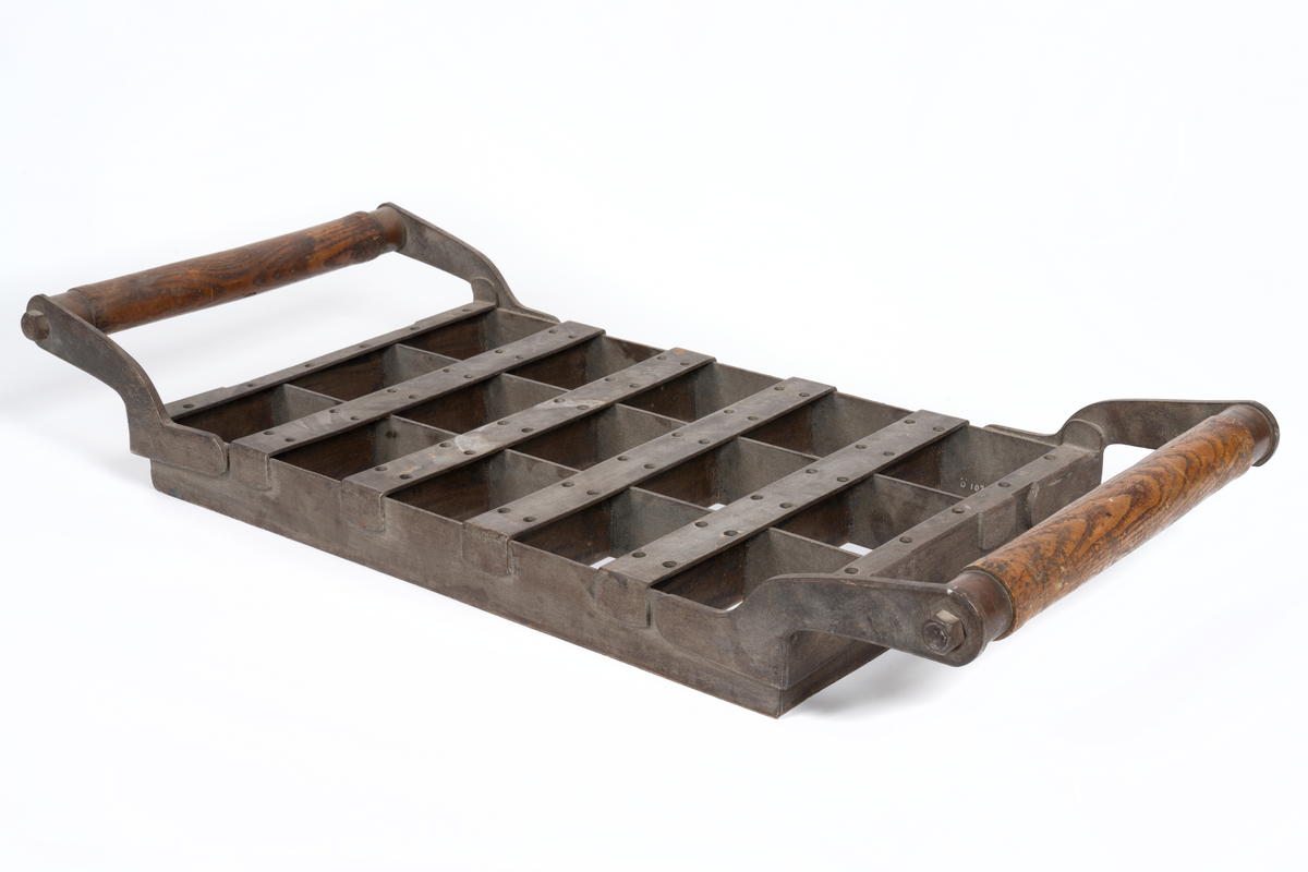 Rektangulär form av järn med två trähandtag längs kortsidorna. Formen är indelad i 15 lika stora rutor. Använd för bakning av skeppsskorpor så kallade knax vid Kronobageriet i Karlskrona.