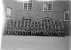 Foto av en pluton meniga i uniform (m/39) framför en kasern (5:e kompaniet) på I 11, Växjö. I mitten av nedre raden syns deras befäl.