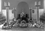 Interiörfoto från (trol. kapellet på Tegnérkyrkogården) inför en begravning. Man ser en mörk kista omgiven av höga kandelabrar och blommor.
