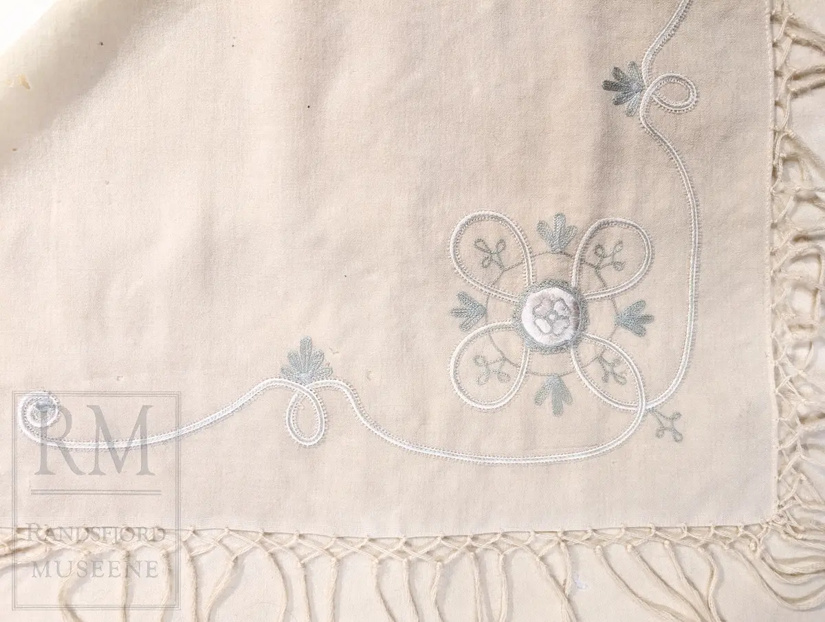 Kvadratisk, tynt ullstoff. Frynser langs alle fire kanter. Brodert motiv i et hjørne, antatt tamburering. 