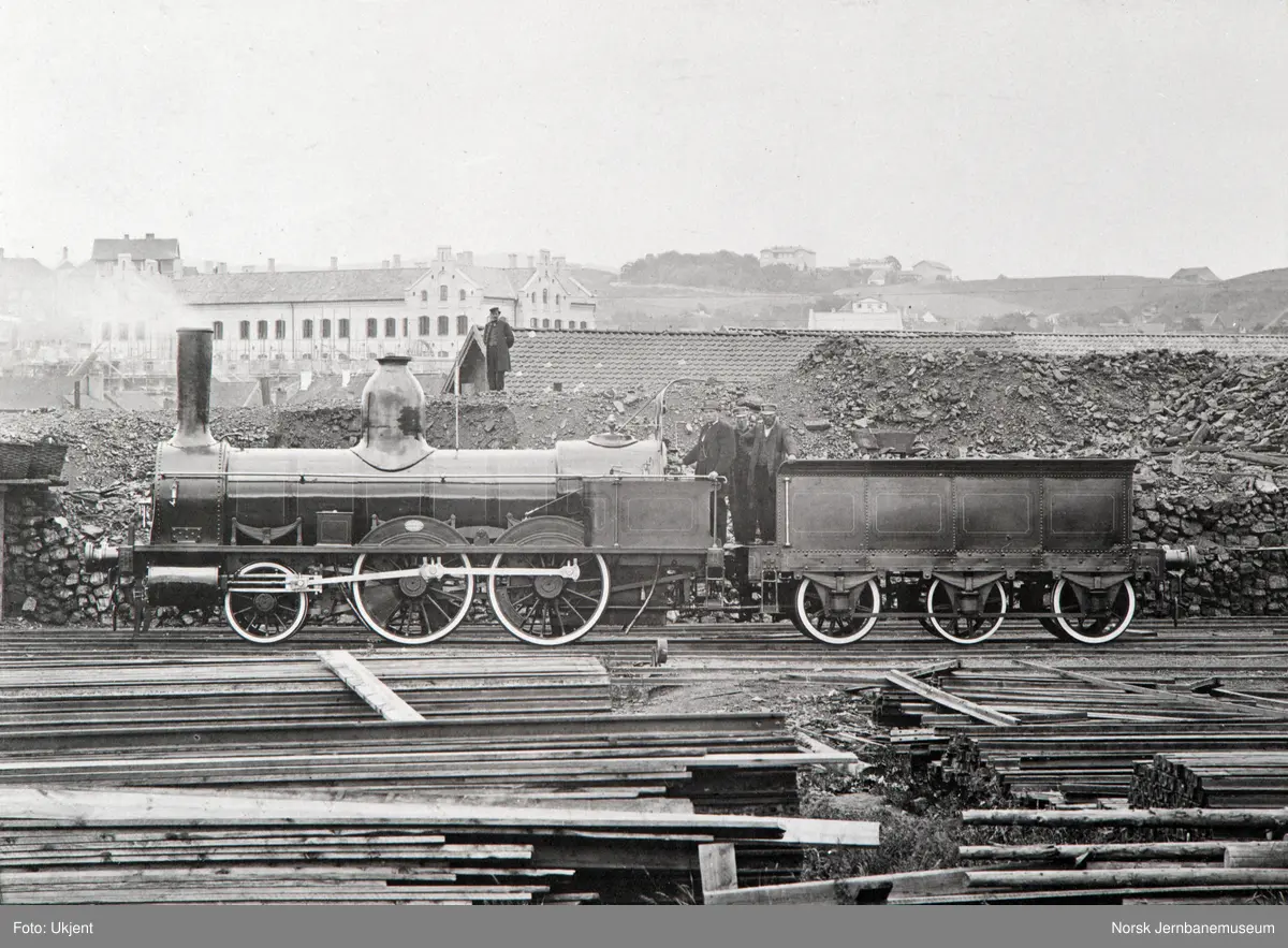 Et av Hovedbanens damplokomotiver nr. 1 - 5 på hovedbanens verkstedtomt, Grundingen 6, senere Oslo gate 3. Den store murbygningen til venstre er Grønlands skole og foran denne er Grønland kirke under oppføring