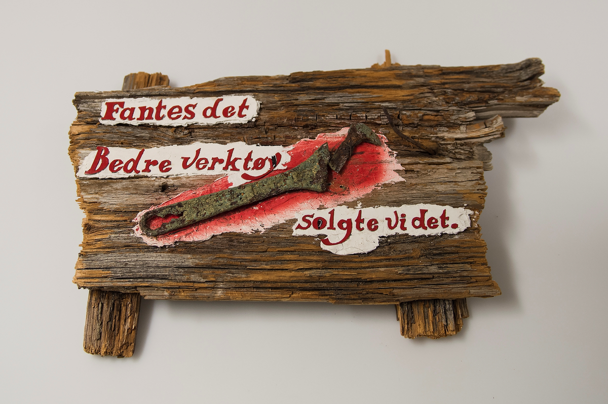 Montasje bestående av treverk fra restene av en gammel palle og en korrodert skiftenøkkel. Bokstaver er skåret ut og limt på de hvitmalte feltene.