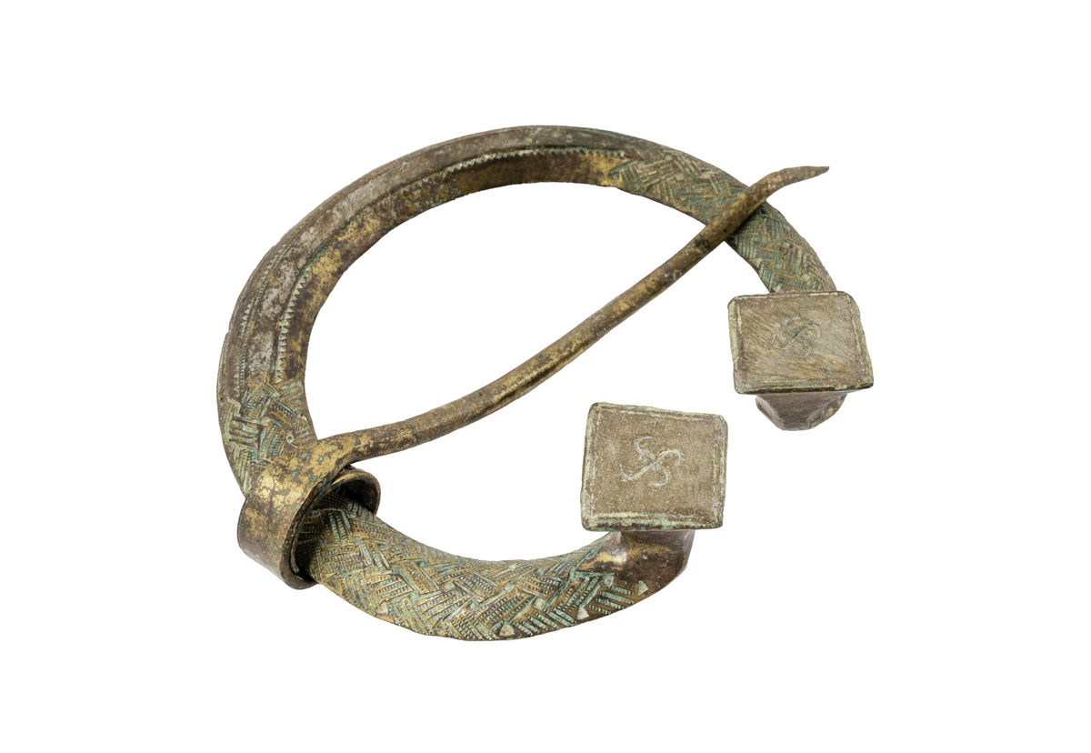 Stort ringspänne med nål. Flätat mönster i relief samt prickat mönster. Båda ändarna formade som uppåtgående fyrkantiga platåer, vardera dekorerad med en svastika i relief. Från Kopparsvik, grav 283.