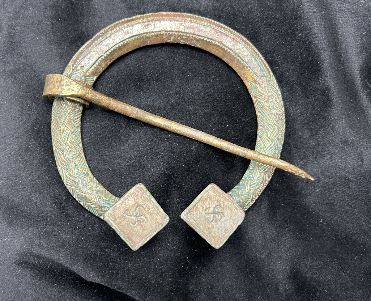 Stort ringspänne med nål. Flätat mönster i relief samt prickat mönster. Båda ändarna formade som uppåtgående fyrkantiga platåer, vardera dekorerad med en svastika i relief. Från Kopparsvik, grav 283.