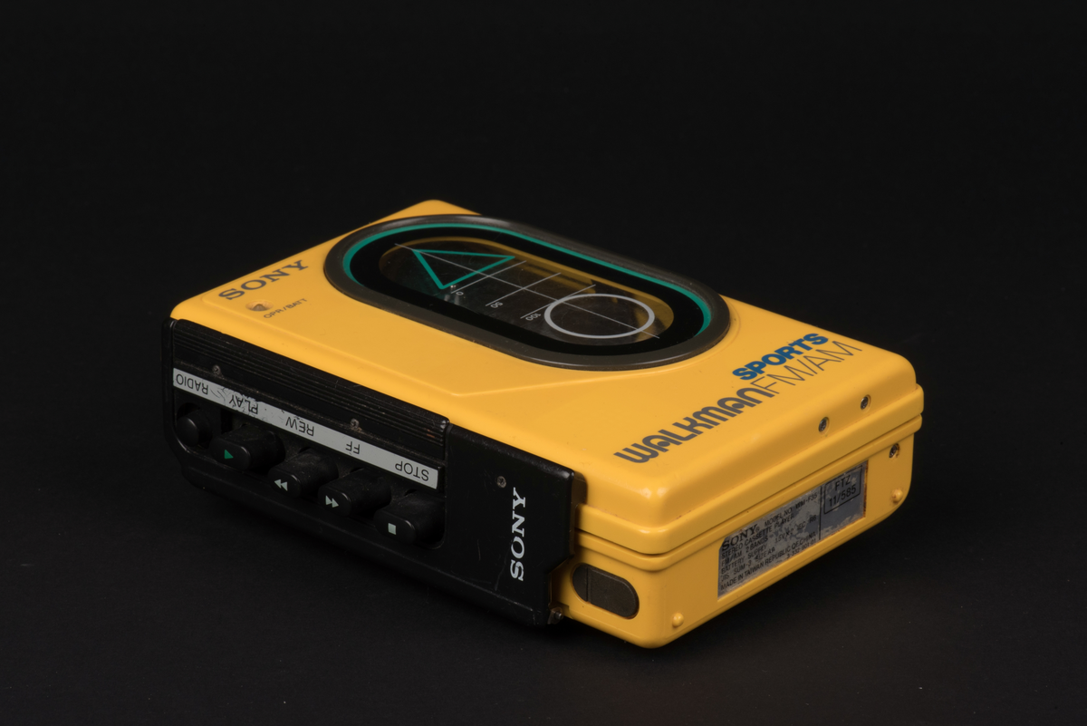 Bärbar kassettbandspelare med radio. Sportmodell som är vatten och stöttålig. Spelaren är tillverkad i gul plast med detaljer i svart och ett fönster i luckan för kassetten.