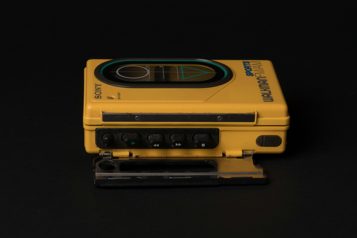 Bärbar kassettbandspelare med radio. Sportmodell som är vatten och stöttålig. Spelaren är tillverkad i gul plast med detaljer i svart och ett fönster i luckan för kassetten.