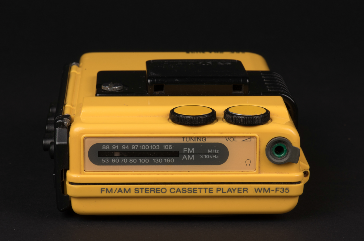 Bärbar kassettbandspelare med radio. Sportmodell som är vatten och stöttålig. Spelaren är tillverkad i gul plast med detaljer i svart och ett fönster i luckan för kassetten.