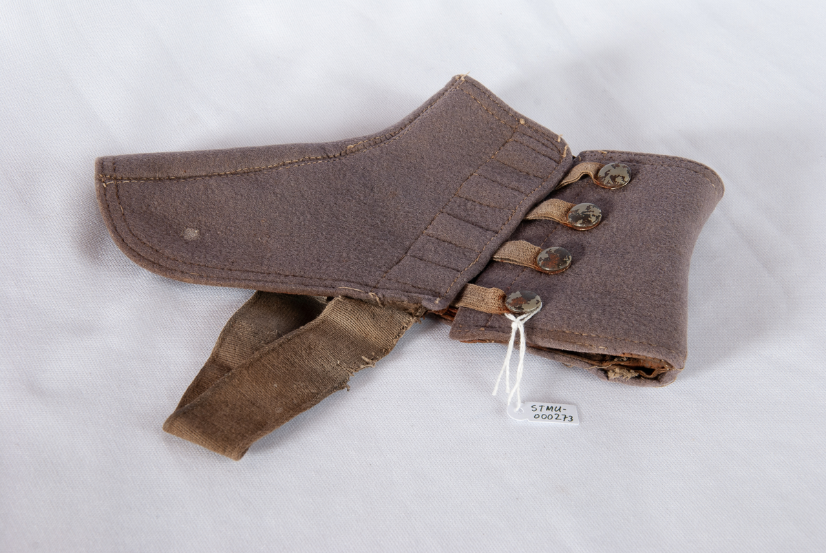 Gamasje. Grå filt. Kneppes på yttersiden med gråmalte trykknapper med strikk. Strikk til feste under skoen. Logo med sort skrift på hvit papir på overkanten. Brun lærkant nederst på innersiden.