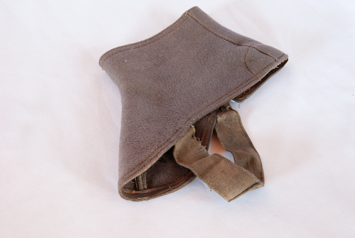Gamasje. Grå filt. Lukkes med 4 gråmalte trykknapper med strikk på yttersiden. Festes med strikk under skoen. Nederkanten er forsterket med lærkant. Logo av papir på overkant med sort skrift.