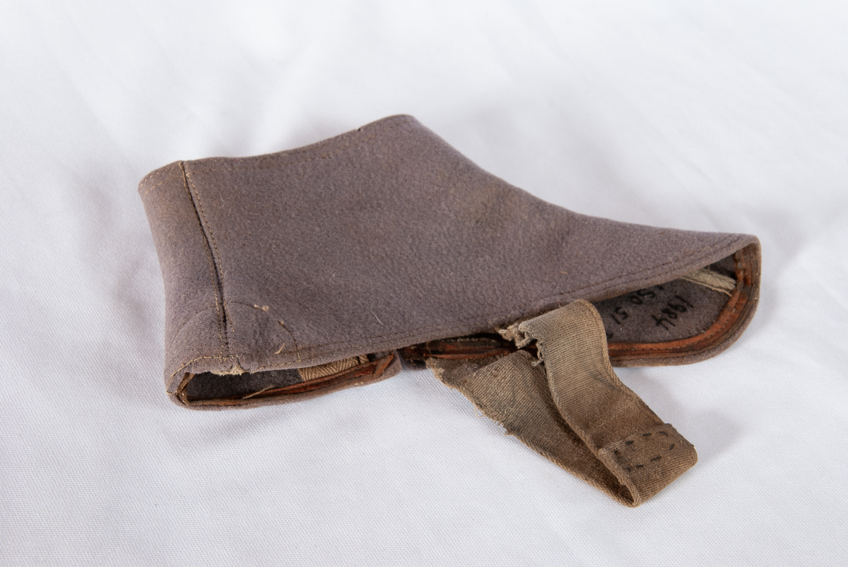 Gamasje. Grå filt. Kneppes på yttersiden med gråmalte trykknapper med strikk. Strikk til feste under skoen. Logo med sort skrift på hvit papir på overkanten. Brun lærkant nederst på innersiden.