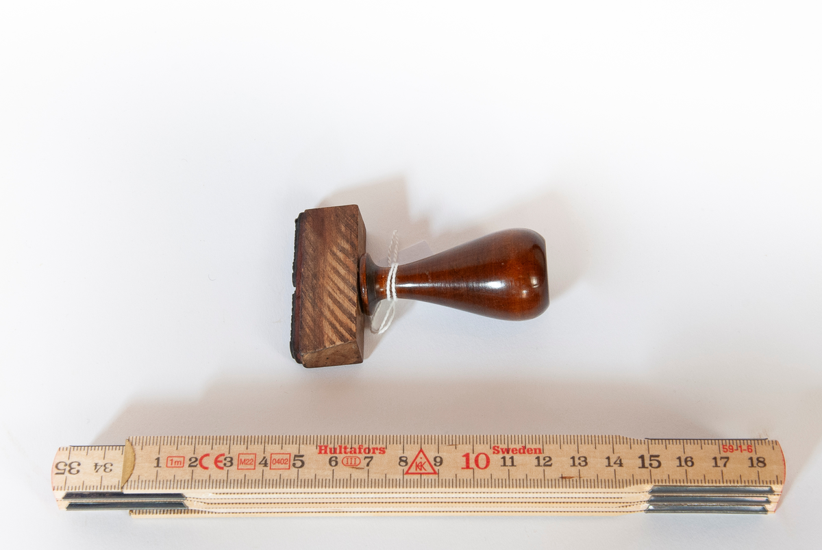 Stempel. Skaft i dreid og lakkert treverk. Dråpeformet med plan ende. På skaftet sirkelformet fabrikkmerke i metall. Til skalftet er festet trestykke med gummiert stempel. 