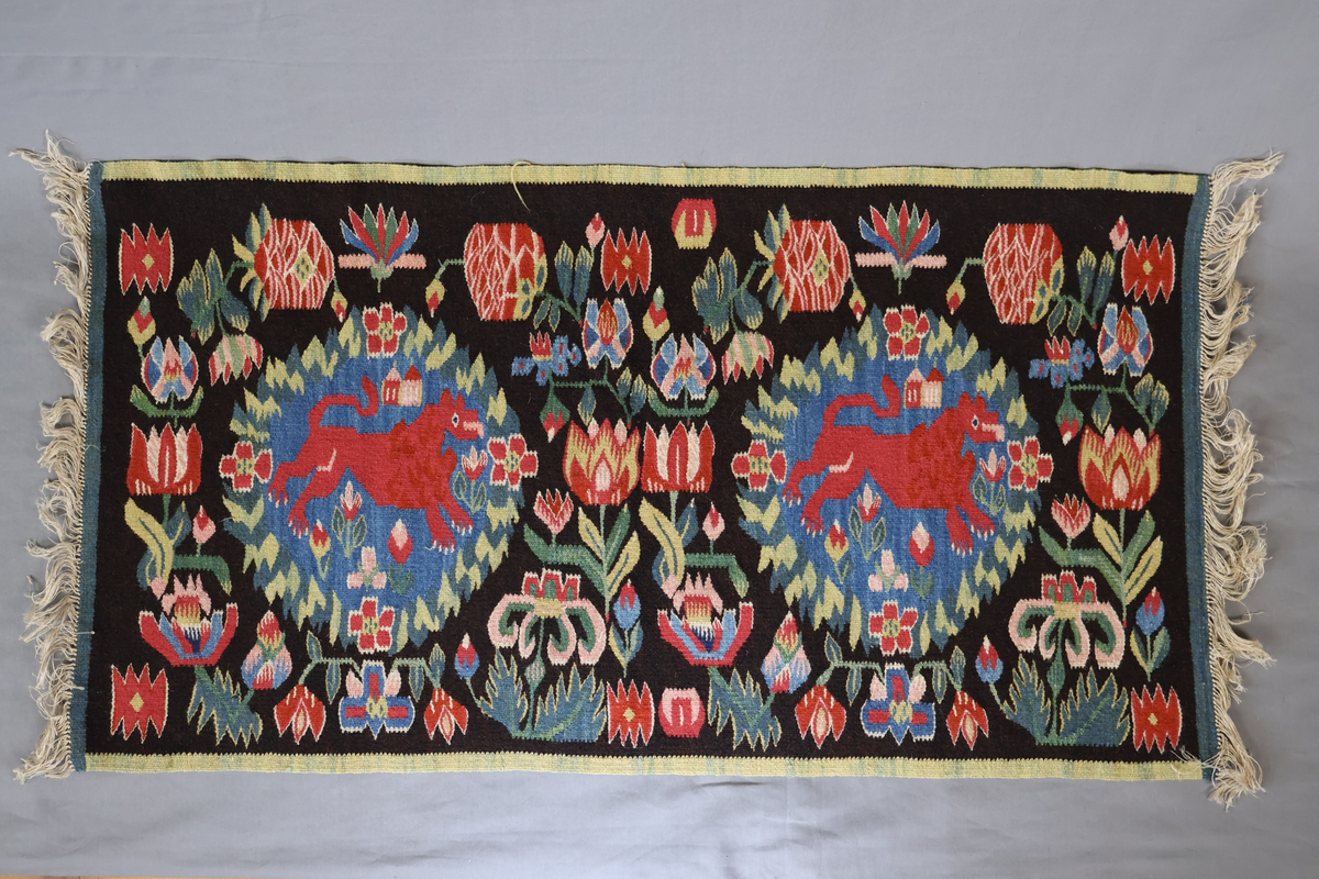 Textil i skånsk flamskvävnadsteknik. Vävd på 1900-talet med en äldre flamskvävnad, inv. nr 303 som förlaga. Denna vävnad är dubbelt så stor/bred som nr. 303 och har två cirklar/kransar med ett rött lejon i varje. Bakgrundsfärgen i kransarna är blå, för övrigt är de två mycket lika. Motiv: rött lejon centralt placerat i en krans av blommor och blad, ca 30 mm i diameter. Tulpaner, liljor mfl blommor spridda runt omkring och i kransen, två små torn i kransen. Ljusgrön, 20 mm bred kant i långsidorna, blågrön, 20 mm bred kant i kortsidorna. Brunsvart botten med mönster i röda, gröna, blå och gula nyanser samt vitt och rosa. Varp i oblekt, 4-trådigt, s-tvinnat lingarn. Inslag i kulört 1-trådigt och 2-trådigt ullgarn, i botten: två trådar svart och en tråd brunt 1-trådigt ullgarn. Inslag i vissa mönsterfigurer: en tråd 2-trådigt och en tråd 1-trådigt ullgarn. Vävnaden är avslutad med knutar och frans av varpen i kortsidorna.