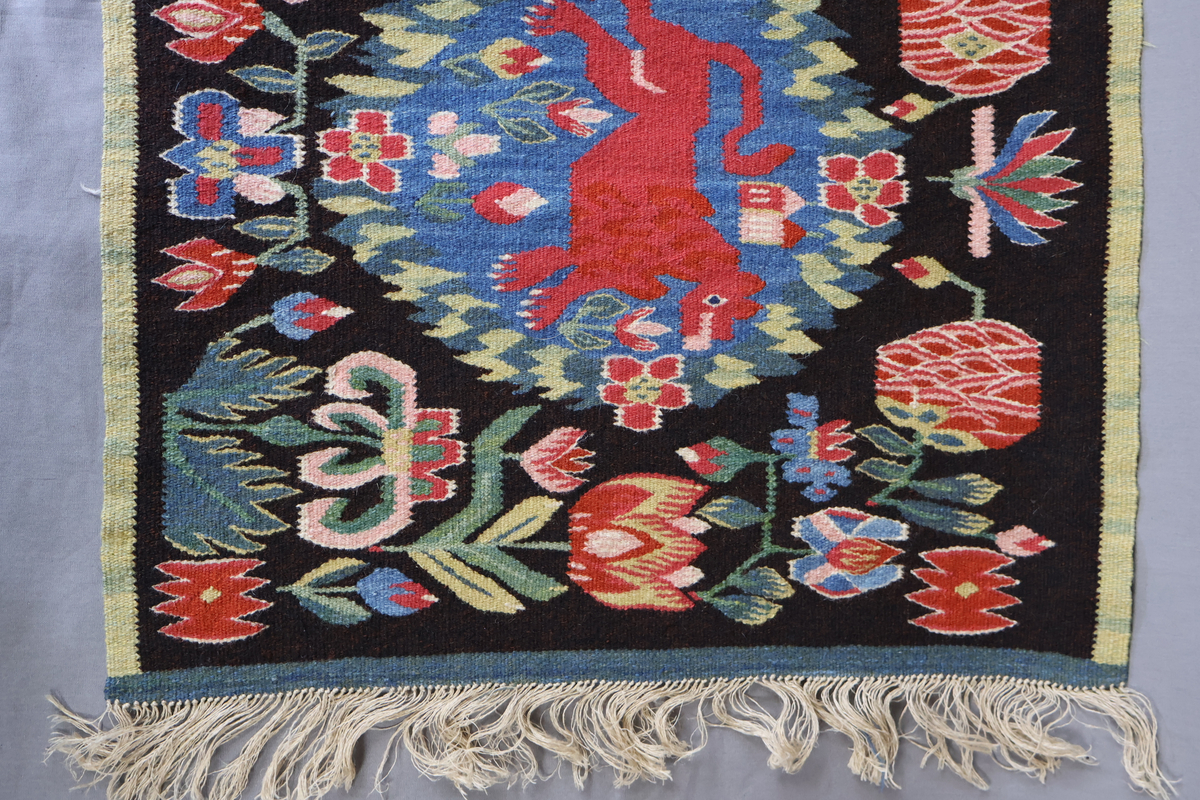 Textil i skånsk flamskvävnadsteknik. Vävd på 1900-talet med en äldre flamskvävnad, inv. nr 303 som förlaga. Denna vävnad är dubbelt så stor/bred som nr. 303 och har två cirklar/kransar med ett rött lejon i varje. Bakgrundsfärgen i kransarna är blå, för övrigt är de två mycket lika. Motiv: rött lejon centralt placerat i en krans av blommor och blad, ca 30 mm i diameter. Tulpaner, liljor mfl blommor spridda runt omkring och i kransen, två små torn i kransen. Ljusgrön, 20 mm bred kant i långsidorna, blågrön, 20 mm bred kant i kortsidorna. Brunsvart botten med mönster i röda, gröna, blå och gula nyanser samt vitt och rosa. Varp i oblekt, 4-trådigt, s-tvinnat lingarn. Inslag i kulört 1-trådigt och 2-trådigt ullgarn, i botten: två trådar svart och en tråd brunt 1-trådigt ullgarn. Inslag i vissa mönsterfigurer: en tråd 2-trådigt och en tråd 1-trådigt ullgarn. Vävnaden är avslutad med knutar och frans av varpen i kortsidorna.