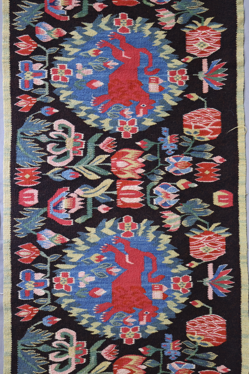 Textil i skånsk flamskvävnadsteknik. Vävd på 1900-talet med en äldre flamskvävnad, inv. nr 303 som förlaga. Denna vävnad är dubbelt så stor/bred som nr. 303 och har två cirklar/kransar med ett rött lejon i varje. Bakgrundsfärgen i kransarna är blå, för övrigt är de två mycket lika. Motiv: rött lejon centralt placerat i en krans av blommor och blad, ca 30 mm i diameter. Tulpaner, liljor mfl blommor spridda runt omkring och i kransen, två små torn i kransen. Ljusgrön, 20 mm bred kant i långsidorna, blågrön, 20 mm bred kant i kortsidorna. Brunsvart botten med mönster i röda, gröna, blå och gula nyanser samt vitt och rosa. Varp i oblekt, 4-trådigt, s-tvinnat lingarn. Inslag i kulört 1-trådigt och 2-trådigt ullgarn, i botten: två trådar svart och en tråd brunt 1-trådigt ullgarn. Inslag i vissa mönsterfigurer: en tråd 2-trådigt och en tråd 1-trådigt ullgarn. Vävnaden är avslutad med knutar och frans av varpen i kortsidorna.