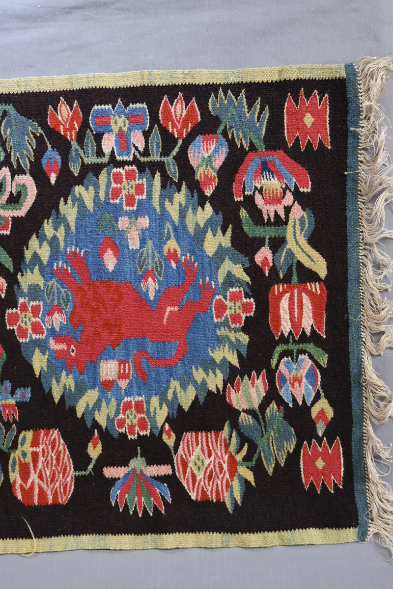 Textil i skånsk flamskvävnadsteknik. Vävd på 1900-talet med en äldre flamskvävnad, inv. nr 303 som förlaga. Denna vävnad är dubbelt så stor/bred som nr. 303 och har två cirklar/kransar med ett rött lejon i varje. Bakgrundsfärgen i kransarna är blå, för övrigt är de två mycket lika. Motiv: rött lejon centralt placerat i en krans av blommor och blad, ca 30 mm i diameter. Tulpaner, liljor mfl blommor spridda runt omkring och i kransen, två små torn i kransen. Ljusgrön, 20 mm bred kant i långsidorna, blågrön, 20 mm bred kant i kortsidorna. Brunsvart botten med mönster i röda, gröna, blå och gula nyanser samt vitt och rosa. Varp i oblekt, 4-trådigt, s-tvinnat lingarn. Inslag i kulört 1-trådigt och 2-trådigt ullgarn, i botten: två trådar svart och en tråd brunt 1-trådigt ullgarn. Inslag i vissa mönsterfigurer: en tråd 2-trådigt och en tråd 1-trådigt ullgarn. Vävnaden är avslutad med knutar och frans av varpen i kortsidorna.