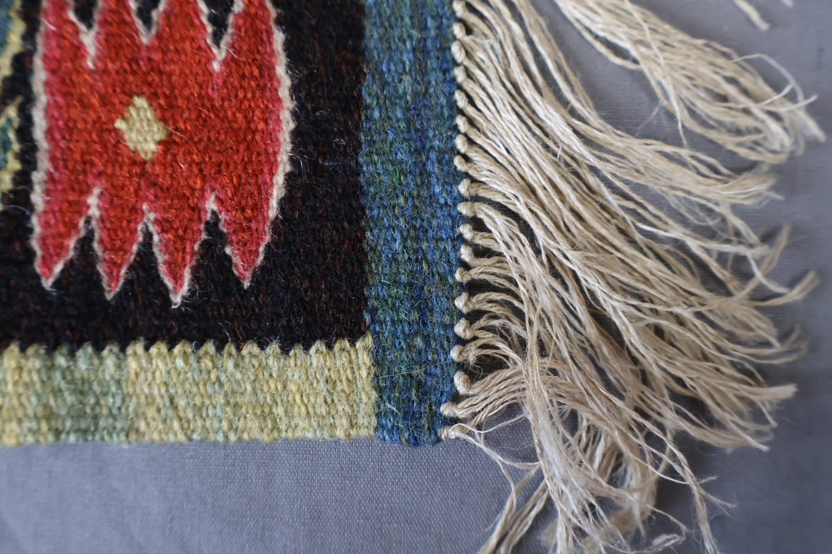Textil i skånsk flamskvävnadsteknik. Vävd på 1900-talet med en äldre flamskvävnad, inv. nr 303 som förlaga. Denna vävnad är dubbelt så stor/bred som nr. 303 och har två cirklar/kransar med ett rött lejon i varje. Bakgrundsfärgen i kransarna är blå, för övrigt är de två mycket lika. Motiv: rött lejon centralt placerat i en krans av blommor och blad, ca 30 mm i diameter. Tulpaner, liljor mfl blommor spridda runt omkring och i kransen, två små torn i kransen. Ljusgrön, 20 mm bred kant i långsidorna, blågrön, 20 mm bred kant i kortsidorna. Brunsvart botten med mönster i röda, gröna, blå och gula nyanser samt vitt och rosa. Varp i oblekt, 4-trådigt, s-tvinnat lingarn. Inslag i kulört 1-trådigt och 2-trådigt ullgarn, i botten: två trådar svart och en tråd brunt 1-trådigt ullgarn. Inslag i vissa mönsterfigurer: en tråd 2-trådigt och en tråd 1-trådigt ullgarn. Vävnaden är avslutad med knutar och frans av varpen i kortsidorna.