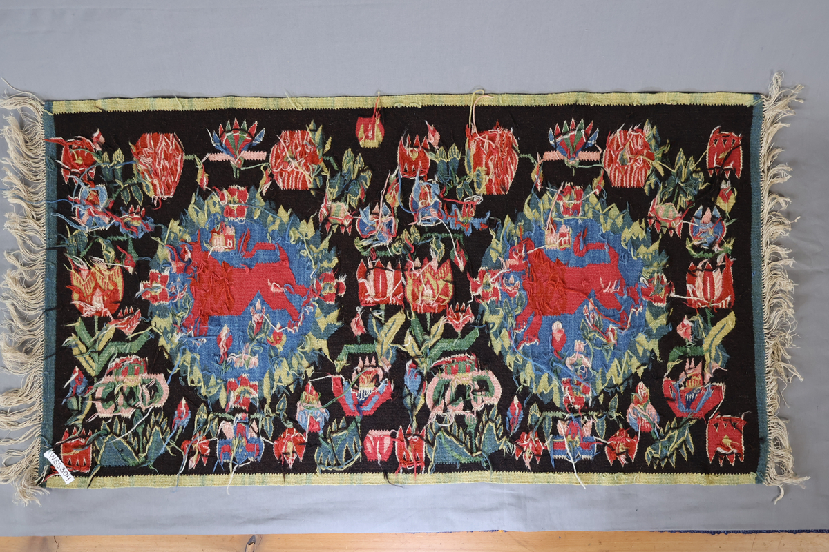 Textil i skånsk flamskvävnadsteknik. Vävd på 1900-talet med en äldre flamskvävnad, inv. nr 303 som förlaga. Denna vävnad är dubbelt så stor/bred som nr. 303 och har två cirklar/kransar med ett rött lejon i varje. Bakgrundsfärgen i kransarna är blå, för övrigt är de två mycket lika. Motiv: rött lejon centralt placerat i en krans av blommor och blad, ca 30 mm i diameter. Tulpaner, liljor mfl blommor spridda runt omkring och i kransen, två små torn i kransen. Ljusgrön, 20 mm bred kant i långsidorna, blågrön, 20 mm bred kant i kortsidorna. Brunsvart botten med mönster i röda, gröna, blå och gula nyanser samt vitt och rosa. Varp i oblekt, 4-trådigt, s-tvinnat lingarn. Inslag i kulört 1-trådigt och 2-trådigt ullgarn, i botten: två trådar svart och en tråd brunt 1-trådigt ullgarn. Inslag i vissa mönsterfigurer: en tråd 2-trådigt och en tråd 1-trådigt ullgarn. Vävnaden är avslutad med knutar och frans av varpen i kortsidorna.