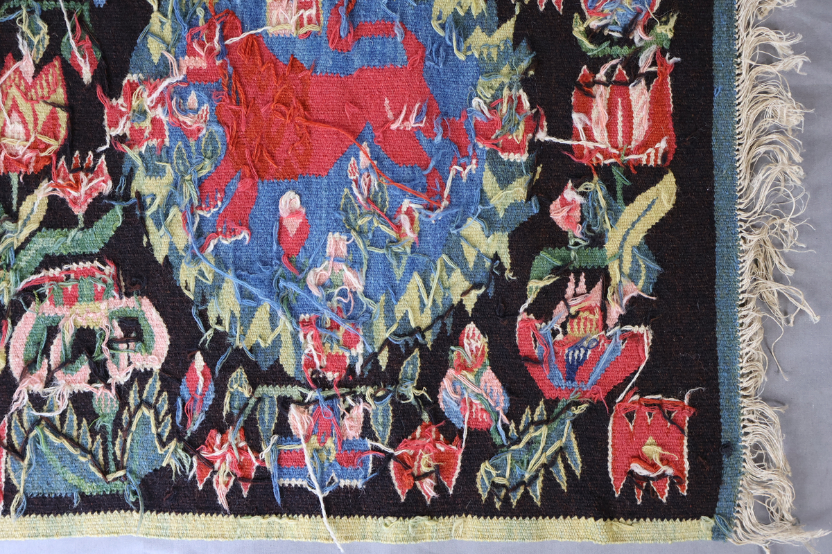 Textil i skånsk flamskvävnadsteknik. Vävd på 1900-talet med en äldre flamskvävnad, inv. nr 303 som förlaga. Denna vävnad är dubbelt så stor/bred som nr. 303 och har två cirklar/kransar med ett rött lejon i varje. Bakgrundsfärgen i kransarna är blå, för övrigt är de två mycket lika. Motiv: rött lejon centralt placerat i en krans av blommor och blad, ca 30 mm i diameter. Tulpaner, liljor mfl blommor spridda runt omkring och i kransen, två små torn i kransen. Ljusgrön, 20 mm bred kant i långsidorna, blågrön, 20 mm bred kant i kortsidorna. Brunsvart botten med mönster i röda, gröna, blå och gula nyanser samt vitt och rosa. Varp i oblekt, 4-trådigt, s-tvinnat lingarn. Inslag i kulört 1-trådigt och 2-trådigt ullgarn, i botten: två trådar svart och en tråd brunt 1-trådigt ullgarn. Inslag i vissa mönsterfigurer: en tråd 2-trådigt och en tråd 1-trådigt ullgarn. Vävnaden är avslutad med knutar och frans av varpen i kortsidorna.