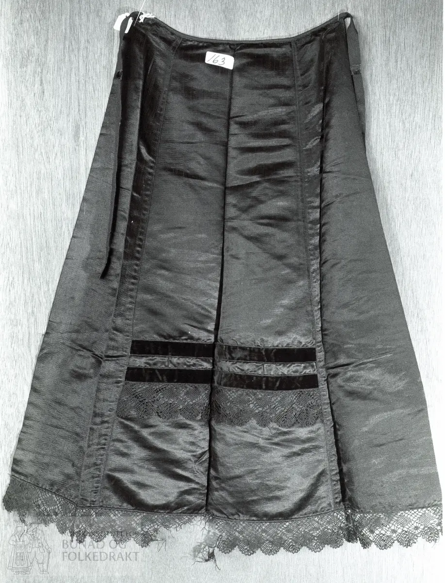 Sannsynleg kjøpt av kremar.  Vart brukt som brurforkle i 1892.  Svart silke, satengvove.  Forkleet er samansydd av 3 skrådde breidder, 4,5 cm brei kniplingsblonde, svart, påsett nederst.  Same type blonde påsett kant på frambreidda, 18 cm.  2 cm breidt fløyelsband sydd på i overkant av blonden.  Same type band er sydd på 1,7 cm ovanfor det andre.  Vidde nede: 74 cm.  Vidde oppe i livet: 30 cm. Høgde:70 cm.  Tillegg blonde: 4,5 cm.