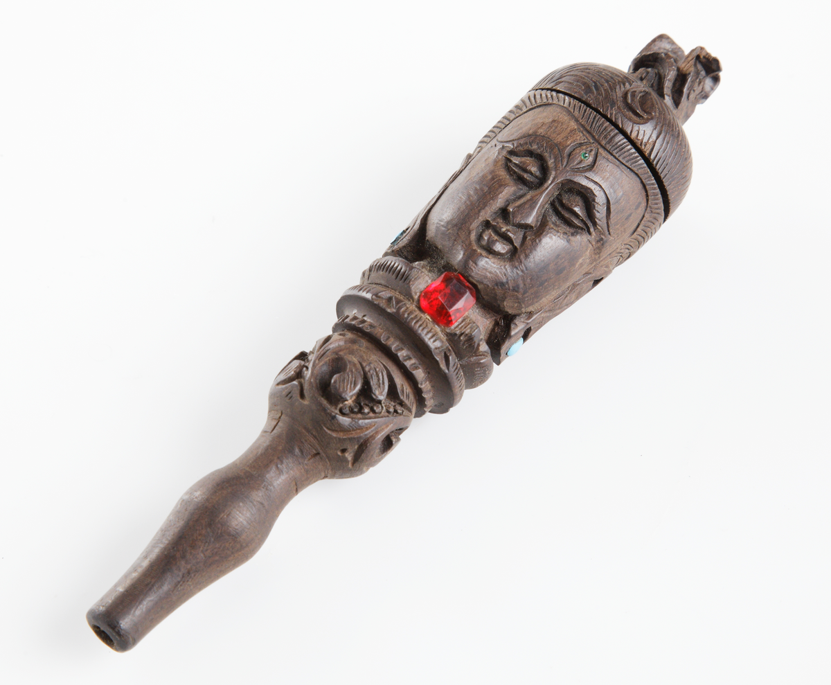 Rak haschpipa i trä med rökutgång framtill på pipan, en så kallad chillum. På holken sitter ett lock placerat. Pipan är svarvad och har ett snidat indiskt ansiktsmotiv runt om. Två ljusblå och en röd glaspärla finns monterade i symboliken.