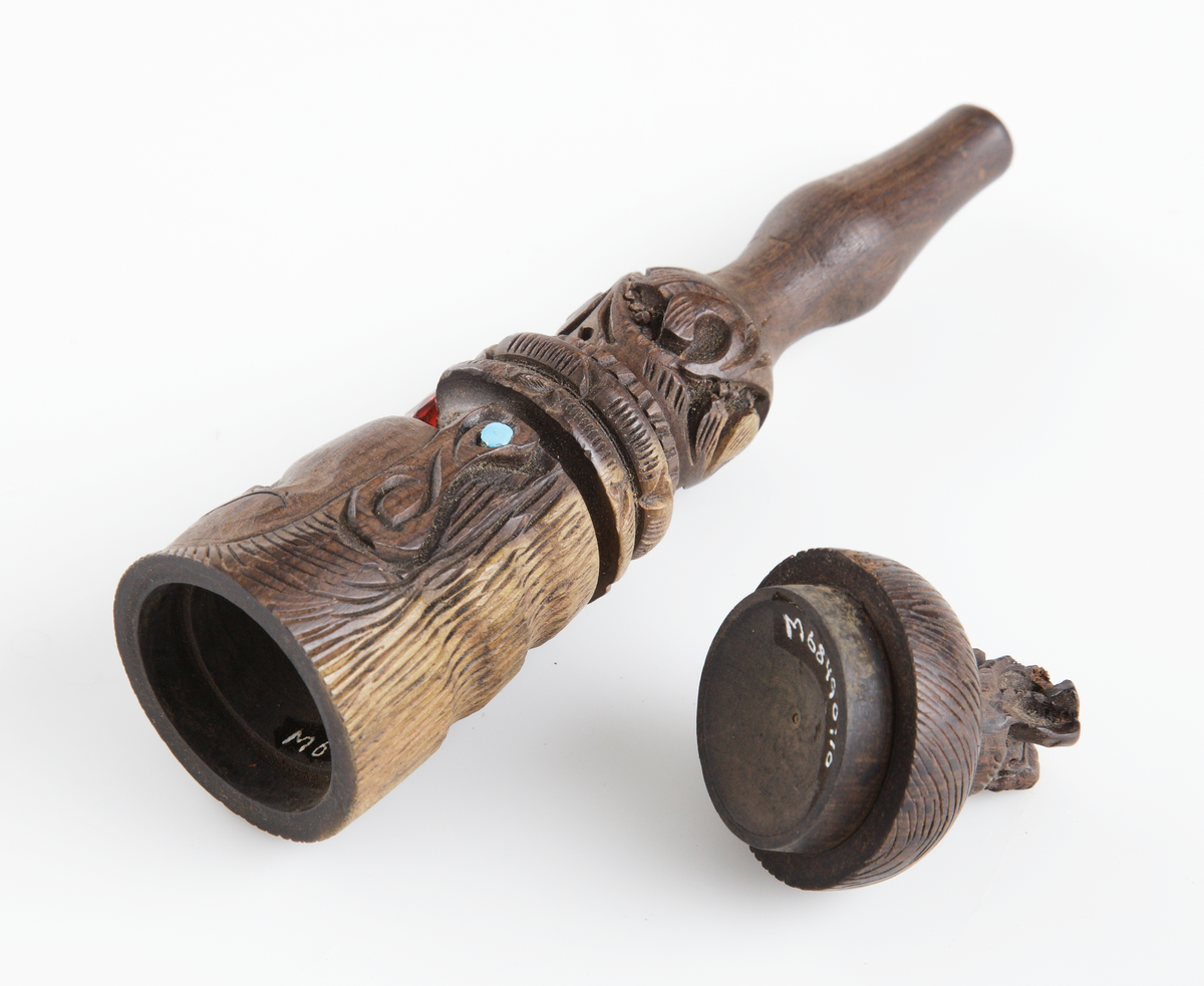 Rak haschpipa i trä med rökutgång framtill på pipan, en så kallad chillum. På holken sitter ett lock placerat. Pipan är svarvad och har ett snidat indiskt ansiktsmotiv runt om. Två ljusblå och en röd glaspärla finns monterade i symboliken.