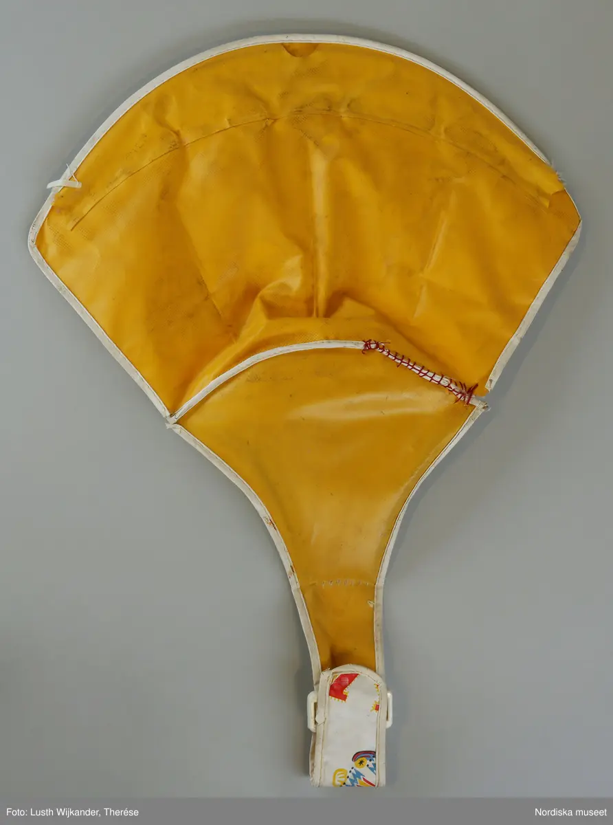 Gångstol (a) med lös sits (b).

a) Gångstol (lära-gå-stol) från 1960-talet, tillverkad i Taiwan. Ställning av metall, fem små hjul och aktivitetsbord av gul hårdplast med aktivitetsknappar i klara färger som barnet kan snurra eller trycka på. Den bärande metallställningen är hopfällbar.

b+) Den avtagbara sitsen av mönstrad pvc-plast är vadderad med skumgummi. Den har sprickor och är lagad med stygn på några ställen. Mönstret avbildar leksaker som teddybjörn, anka, docka och tåg samt bokstäver mot vit botten. Undersidan gul. Sitsen sitter fast vid ställningen genom en bågformad metallten som löper genom  sitsens ryggparti. 

Ihopmontering av sits och bord: Metalltenen skjuts in i överkanten på sitsens ryggparti. Sitsen sätts i hålet på aktivitetsbordet och sitsens metallten fästen i de små hålen på sidorna av hålet i aktivitetsbordet samt i ett fyrkantigt hål baktill på aktivitetsborden med hjälp av ett metallbleck som omsluter metalltenen.  Den smala framdelen/fliken av sitsen träs runt metallpinnen som sitter på aktivitetsbordets undersida och fästes genom plastspännet.

Anm. Nött och delvis något solkig. Sitsen är lagad med stygn på ett par ställen.
/Karin Dern 2012-11-15