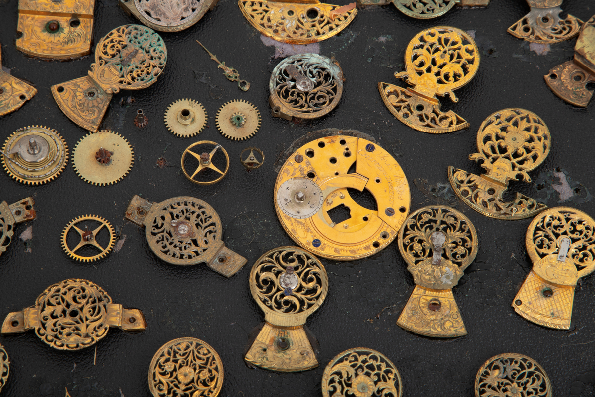 Samling av urdeler, cirka 50 stykk, de fleste av delene er klober. Delene er limt på en papplate som er festet i en trekasse med hengslet glasslokk.