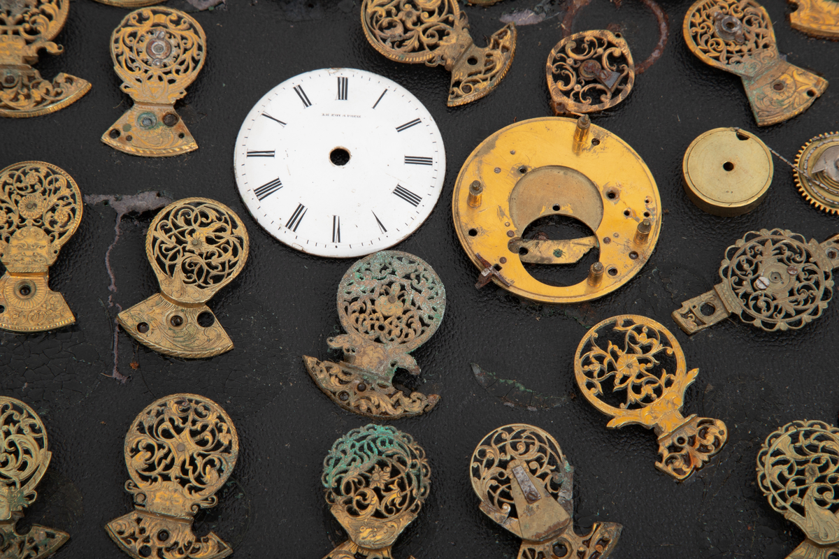 Samling av urdeler, cirka 50 stykk, de fleste av delene er klober. Delene er limt på en papplate som er festet i en trekasse med hengslet glasslokk.