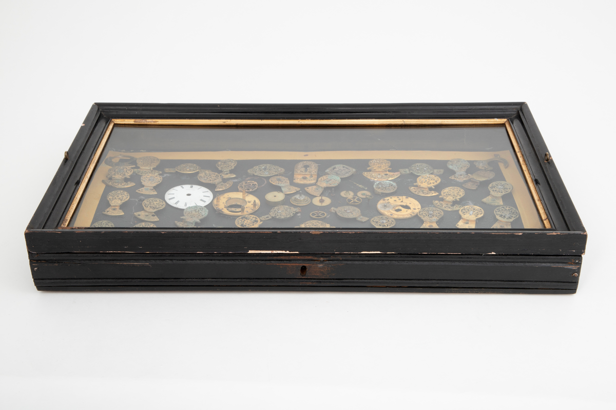 Samling av urdeler, cirka 50 stykk, de fleste av delene er klober. Delene er limt på en papplate som er festet i en trekasse med hengslet glasslokk.