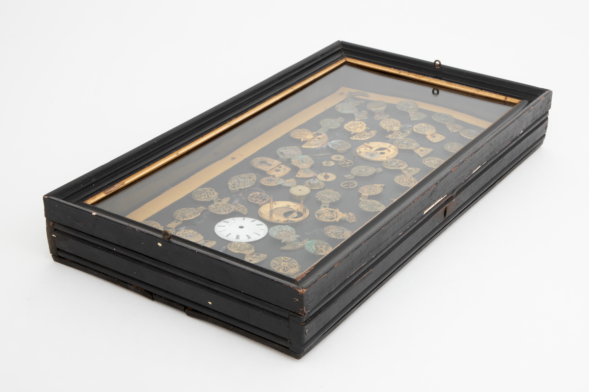 Samling av urdeler, cirka 50 stykk, de fleste av delene er klober. Delene er limt på en papplate som er festet i en trekasse med hengslet glasslokk.