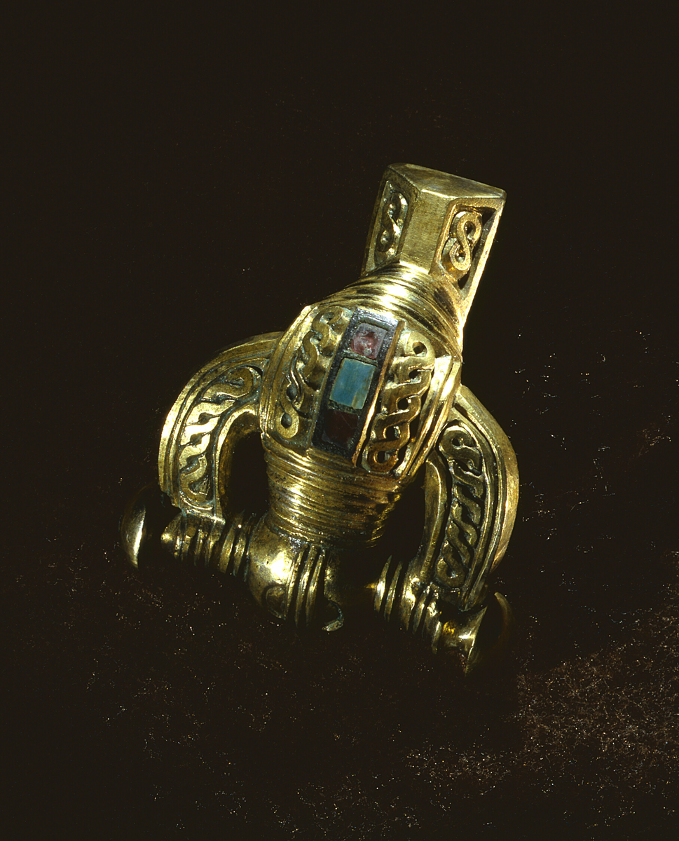 Spänne, så kallat "kräftspänne", från kvinnograv i Lau socken.

På bygelns krön finns tre infällda stenar i cloisonnéteknik, två röda och en turkosfärgad. De röda stenarna består sannolikt av halvädelstenen granat och den turkosa är en bit av horn eller ben. Den ljusblå färgen erhålls troligtvis av låga halter av kopparsalter på ytan.

Spännet är dekorerat med bandflätor av typ liggande åttor.

Någon motsvarighet till det förgyllda spännet finns inte på Gotland och från svenska fastlandet finns endast några liknande exemplar.