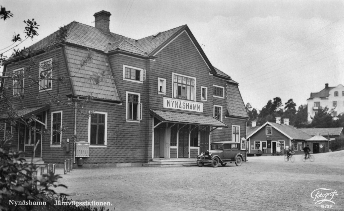Nynäshamns järnvägsstation. Framför stationen står en bil parkerad, några barn cyklar förbi. I bakgrunden syns Antons Styréns Speceri- och diversehandel. 

Järnväg, kommunikation.