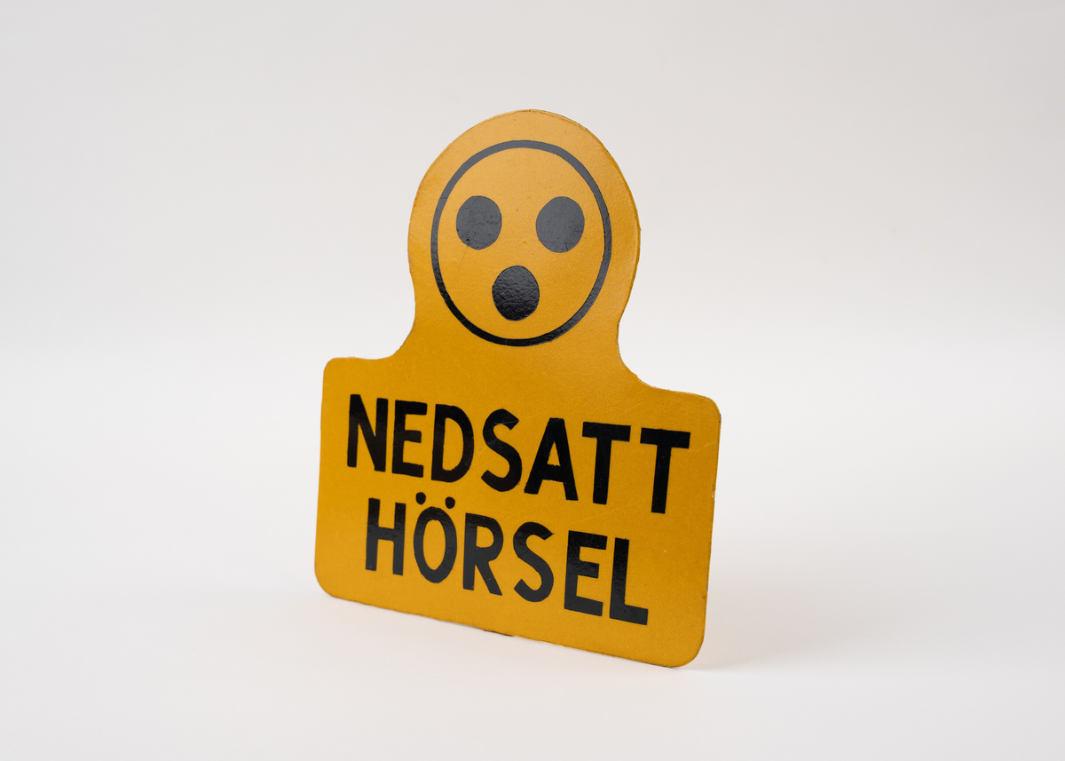 Gul skylt i metall med svart text: "NEDSATT HÖRSEL". Ovanför texten finns den välkända symbolen med tre svarta prickar i en cirkel – en symbol som ofta används för att signalera hörselnedsättning. Skylten är formad med en avrundad topp och utbuktningar i sidorna. På baksidan finns ett fastsvetsat rörfäste som möjliggör montering, t.ex. på en annat fäste.

Skylten användes för att uppmärksamma omgivningen på att personen som har nedsatt hörsel, och kan behöva särskild hänsyn vid kommunikation. Den har använts i offentliga miljöer. 