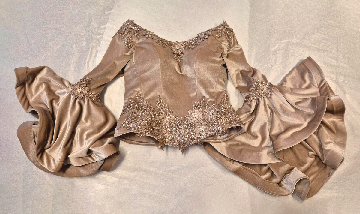 Strukturerad överdel uppsydd i rosa-beige sammetstyg och champangefärgade applikationer samt stenar.  Applikationerna är i form av blommor och löv. Fastklistrade på tyget. De är placerade runt den runda ringningen, lätt nedåt vid axeln samt nedre delen av plagget. Torson är uppdelad i 10 tygpaneler ihopsydda med raksöm, fodrad med ett beige tyg (möjligen baksidan av sammetstyget). Stängda kanaler på insidan för korsettbening.16 stycken totalt.  Nedre kanten av överdelen är benad. Inbyggda kupor. Korsettstängning på baksidan med extra tygstycke för att dölja huden under. Stängningen består av 10 stycken tyg öglor på vardera sidan av överdelen och ett tillhörande band.  

Ärmarna är fästa till överdelen med zick-zack söm. Raka, smala ärmar till armbågen, därefter öppnas de upp till en vid fallande form. Ofodrade vid den smala delen. Den vida delen är fodrad med samma sammetstyg som yttertyget. 
Dom smala delarna är ett tygstycke vardera. De vida delarna är ett separat tygstycke vardera och fästa till resterande ärm med raksöm, lätt veckade. 
Ärmarna avslutas med en fållad kant som innehåller ett stabiliserande mellanlägg. Applikationerna samma som ovan men placerade kring armbågen - där den smala delen av ärmen möter den vida delen. 



