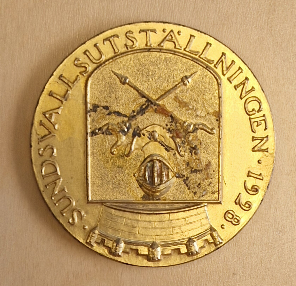 Medalj av metall. Framsidan: ett krönt stadsvapen, Sundsvallsutställningen 1928. Baksidan: Inom en krans: Rudolf Granberg Sundsvall och svårlästa stämplar: ?, 5809, kattfot, 33 eller BB. Gåva av Owe Walter.

Anm: Under högsommaren 1928, den 21 juni till den 29 juli anordnades en sommarutställning i Sundsvall. Ungefär 850 utställare från de mest varierade verksamheter lockade till sig mängder med besökare.

Året innan, 1927, hade Ostkustbanans räls anlagts och tågen till Sundsvall hade börjat gå längs kusten. Det underlättade kommunikationerna betänkligt, och var en orsak till att utställningen förlades hit. Olof Ahlström, Sundsvalls Tidnings fd chefredaktör var initiativtagare och generalsekreterare för mässan.

Alltihop invigdes den 21 juni av kronprinsen Gustaf Adolf och hans hustru Louise Mountbatten. Redan den första dagen löste 8000 personer in sig på området och det var festyra i staden.

Utställningen låg inne på de olika skolgårdarna längs Skolhusallén och fortsatte en bit upp mot Södermalm där ett nöjesfält slagit upp sina karuseller.

En hel del nya byggnader fick uppföras, bland annat en stor maskinhall och en 112 meter lång automobilhall på läroverksplan. En viadukt över järnvägen och en restaurant på folkskoleplanen samt en byggnad för hästutställningen byggdes också upp enbart för utställningen. Att det fanns stora hallar både för bilar och hästar visar på den brytningstid som tjugotalet innebar när det gäller den allt mer utbredda motoriseringen.

På nöjesfältet fanns attraktioner som en dansbana och ett dåtidens lustiga hus med rörliga golv, rullande trottoarer och skrattspeglar. Ett spektakulärt högt torn i sex våningar med rutschkanor i spiral reste sig också högt över åskådarna på marken.

Utställningen som pågick i en månad var den största i sitt slag som då hållits i Norrland och lockade utställare inom näringar som exempelvis industri, hantverk, livsmedel, jordbruk, konst och turism. Föreningar som Skidfrämjandet var också på plats, mitt i sommarvärmen, för att marknadsföra Storlien som skidort.

I samband med utställningen anordnades även två motortävlingar, en mer professionell tävling mellan Stockholm och Sundsvall och en "turisttävling" mellan Falun och Sundsvall. Fler tävlingar, i form av spinntävlingar och annat hantverk var också populärt och utställningar av både frön och konst var viktiga delar.
Källa: Sundsvalls Tidning 8 september 2013 