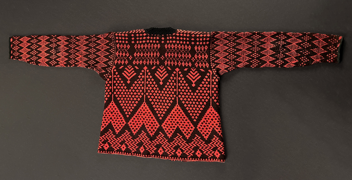Herrtröja, av rött och svart ullgarn. Rundstickad bål och ärmar, öppen fram, knäpps med sex svarta knappar. Öppningen fram och ärmarna skodda med band av svart diagonalvävd ylle. I halsen en stickad krage i två lager av svart ylle. Slät insida, hålmönstrad utsida. Skoningen och kragen troligen sekundära.