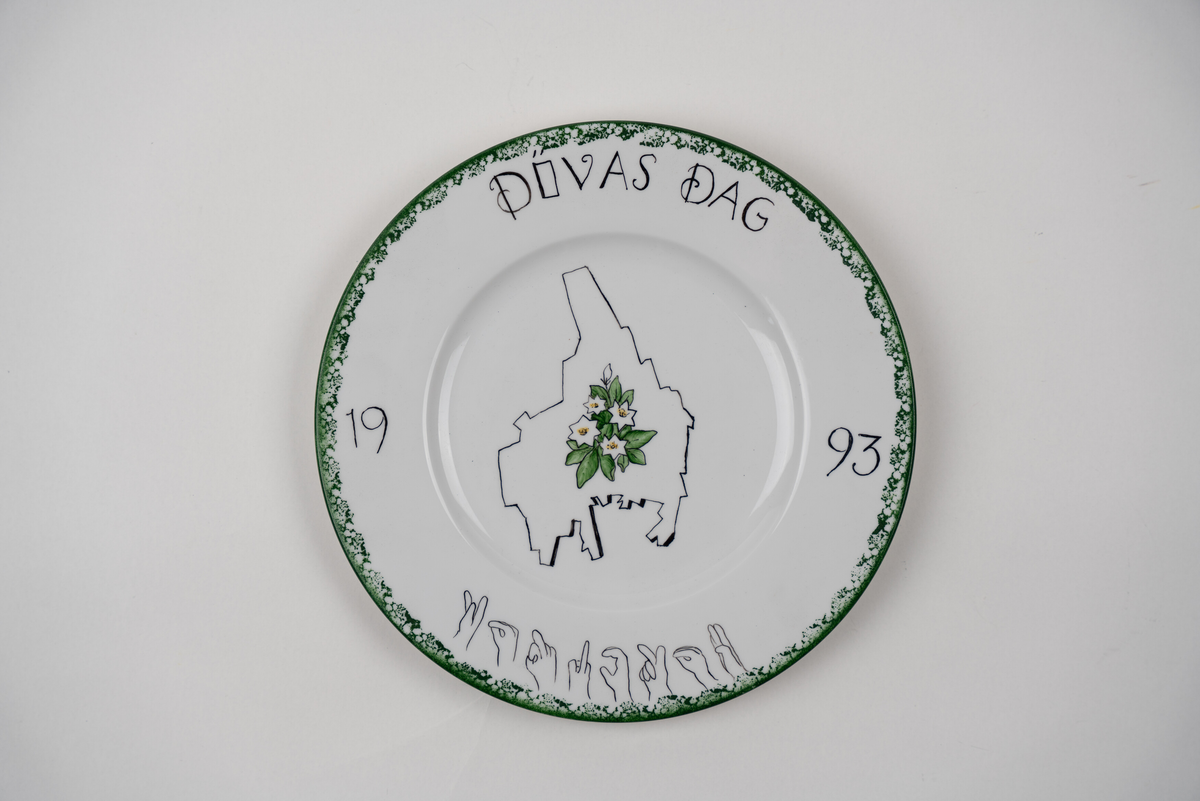 Väggtallrik, handmålad, med illustration av konturen av Värmlands lanskap, landskapsblomman skogsstjärna. Text: "Dövas dag 1993", Karlstad är skrivet med handformerna i svenska handalfabetet. Handmålad av Siv Karlsson. 


