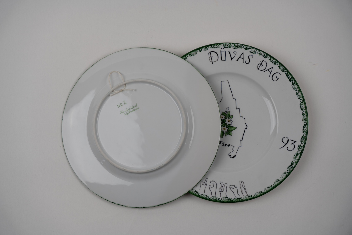 Väggtallrik, handmålad, med illustration av konturen av Värmlands lanskap, landskapsblomman skogsstjärna. Text: "Dövas dag 1993", Karlstad är skrivet med handformerna i svenska handalfabetet. Handmålad av Siv Karlsson. 


