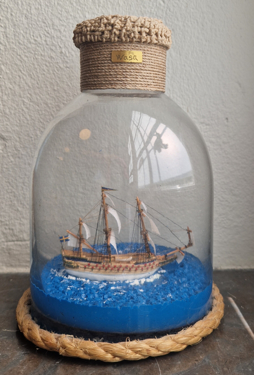 Stående flaskskepp, kantat med rep nedtill. En liten metallskylt på båda sidor "Wasa". Tillverkare Per Hugo Hedebrand. Flaskan är förseglad med mässingsplakett och makramé, plakett med inristad monogramsignering, A P H + ett ankare. Gåva av Ing Marie Hedebrand, Sundsvall. 